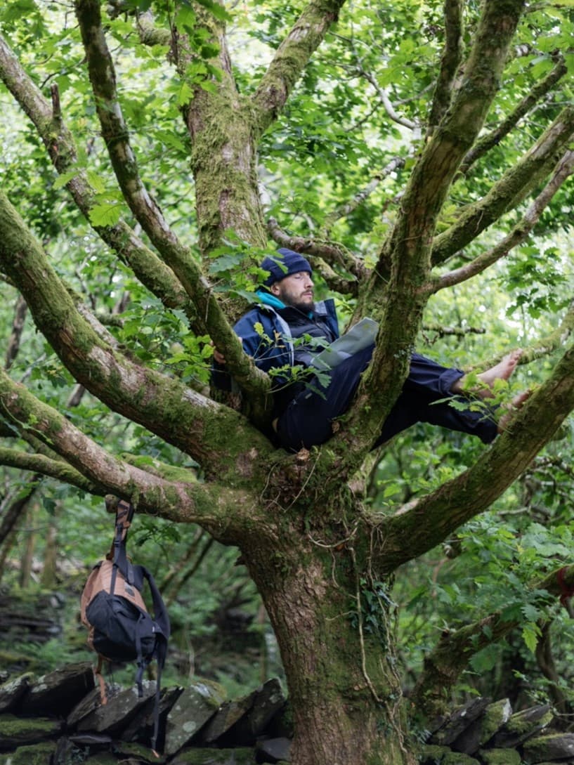 A person in winter clothes sat barefoot in a tree, backpack hanging from a branch.