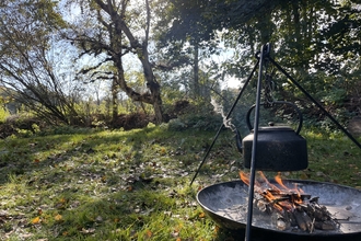 A kettle hanging over the flames over a campfire, with autumn trees behind