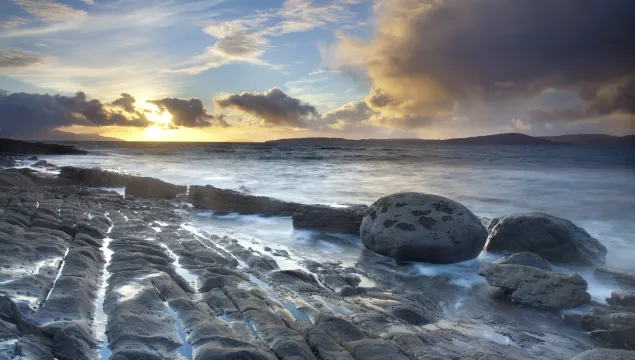 A coastal landscape, with the sea gently lapping at smooth rocks as the sun sets behind scattered clouds