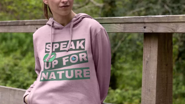 Speak up for nature hoodie worn by a person who's leaning on a fence post in a wooded area