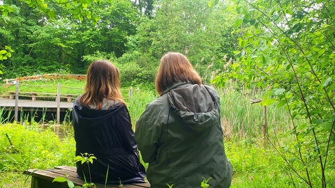 Two people looking away from the camera, sat on a bench surrounded by green leaves
