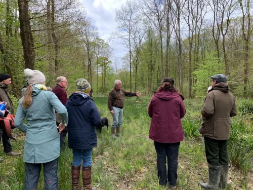 A group of people in raincoats surround a man talking and pointing to the environment around them.