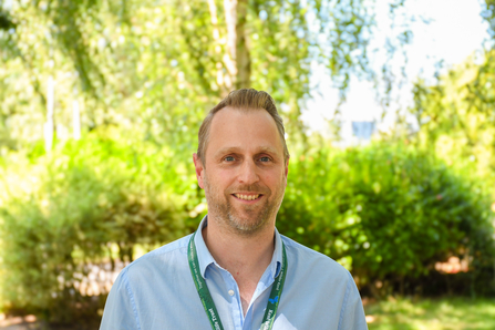 Tom Woolgar, trustee, in front of green trees and bushes.